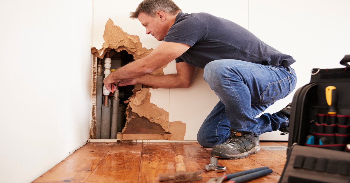 A man inside of a home wearing a shirt and jeans kneeling in front of a pipe in the wall making repairs.