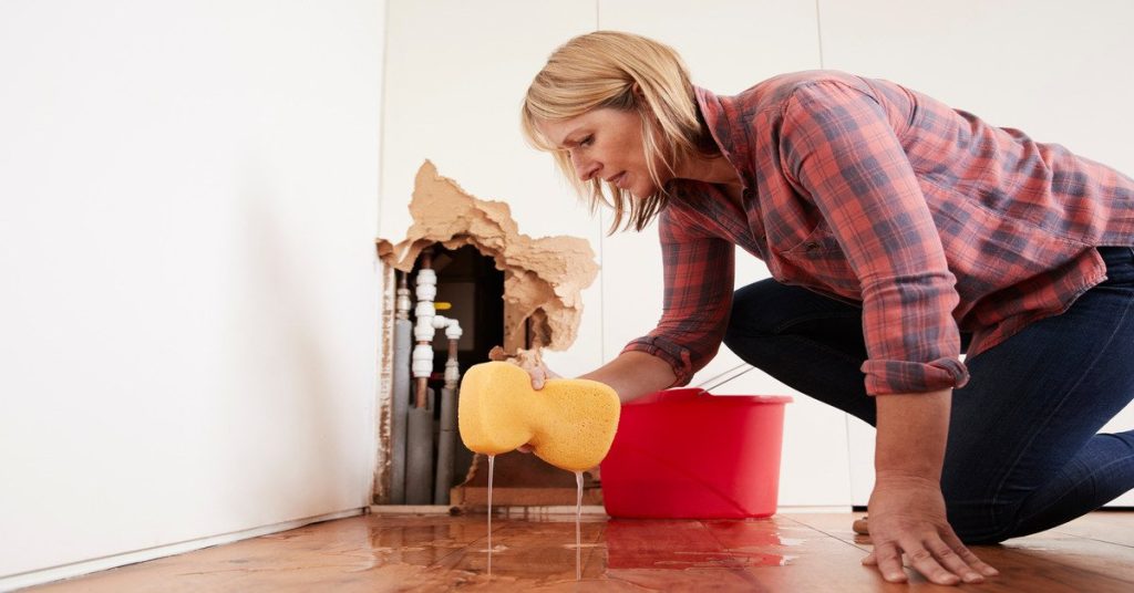 A woman wearing a flannel shirt and jeans kneeling on the floor in her home cleaning up water from burst pipes.